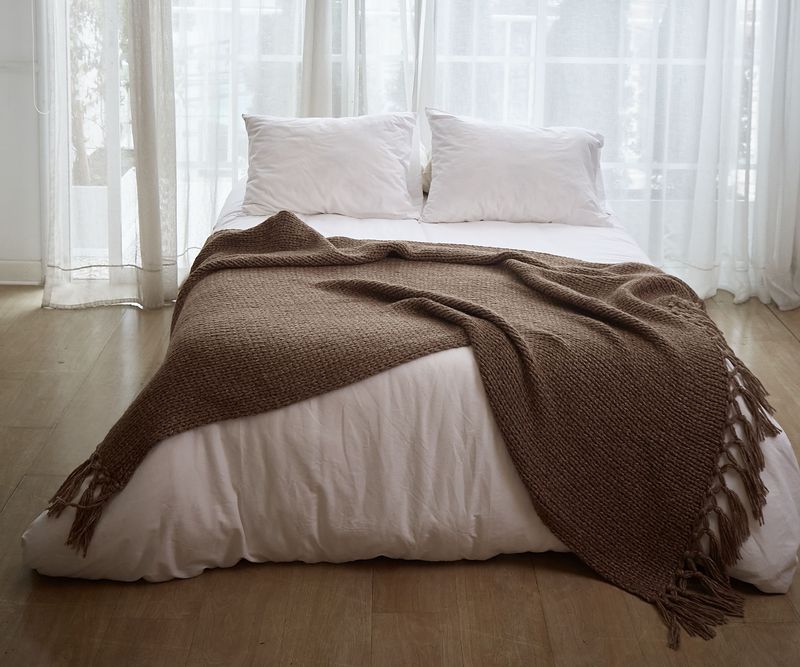 A cocoa brown chunky-knit throw with knotted tassels, draped over a white bed in front of sheer white curtains